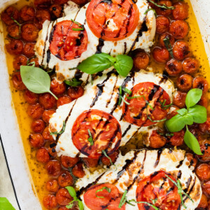 Three chicken breasts covered in mozzarella cheese, tomato slices, and balsamic glaze in a rectangular baking dish with cherry tomatoes and fresh basil.