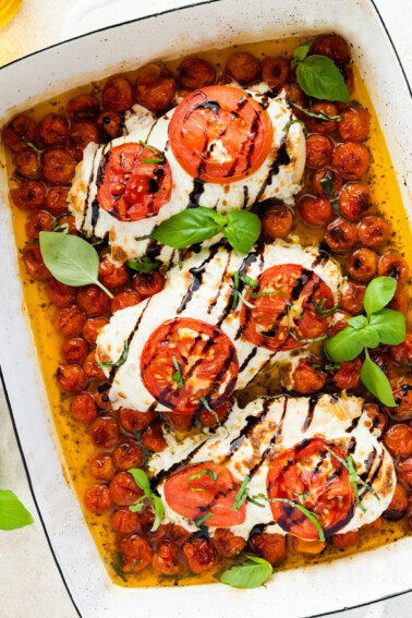 Three chicken breasts covered in mozzarella cheese, tomato slices, and balsamic glaze in a rectangular baking dish with cherry tomatoes and fresh basil.