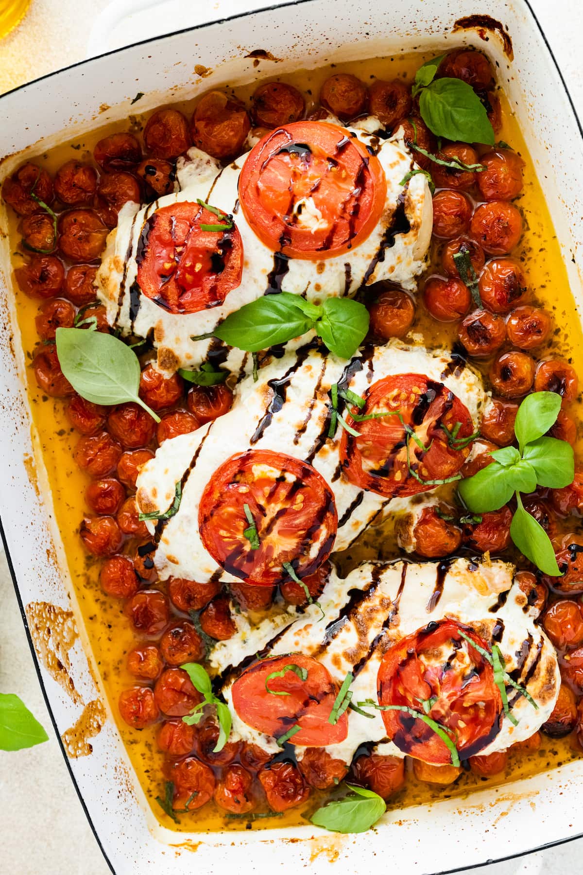 Three chicken breasts covered in mozzarella cheese, tomato slices, and balsamic glaze in a rectangular baking dish with cherry tomatoes and fresh basil.