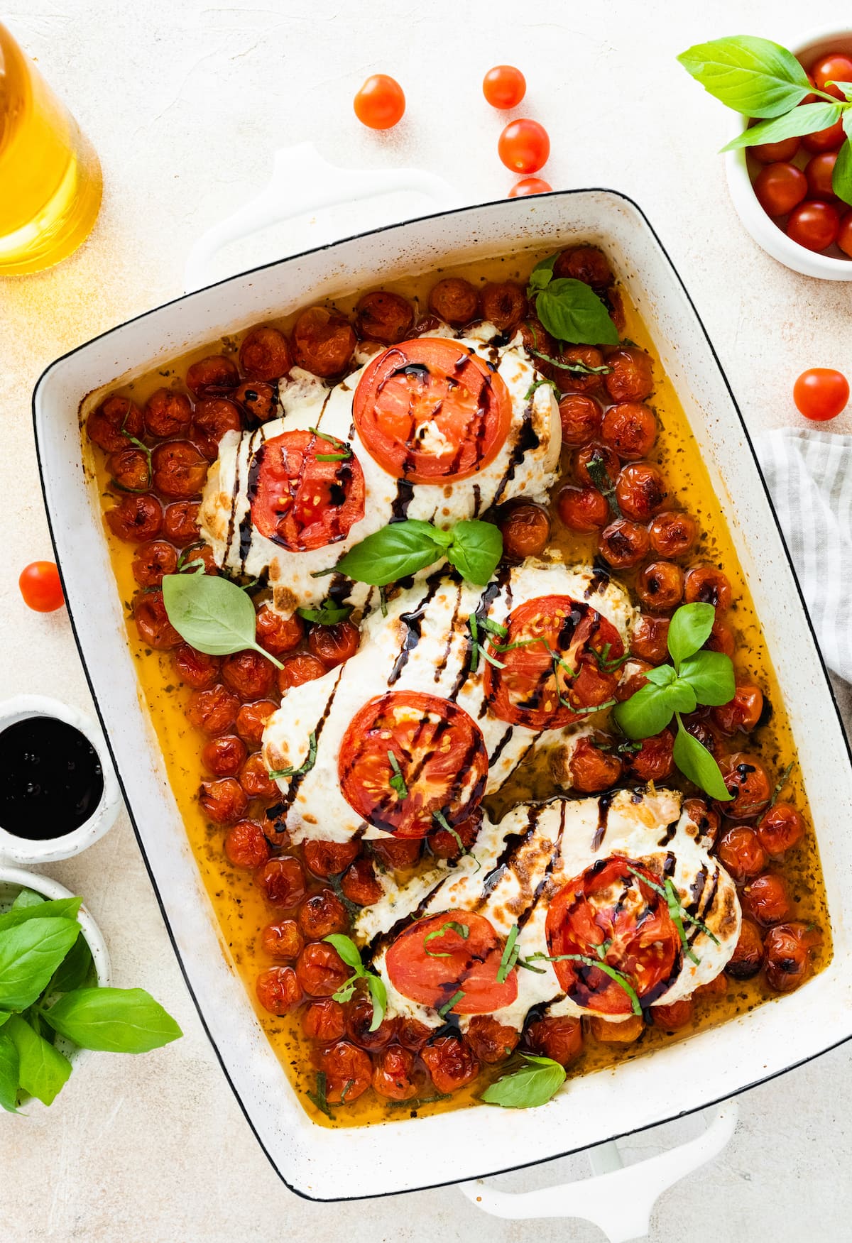 Three chicken breasts covered in mozzarella cheese, tomato slices, and balsamic glaze in a rectangular baking dish with cherry tomatoes and fresh basil.