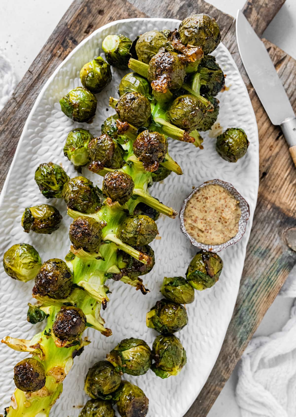 Roasted Brussels Sprouts on the Stalk on a large serving plate on top of a cutting board near a small bowl of mustard and a knife.