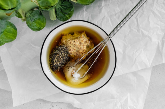 A small bowl with a small whisk containing mustard, salt, pepper, oil, and honey.