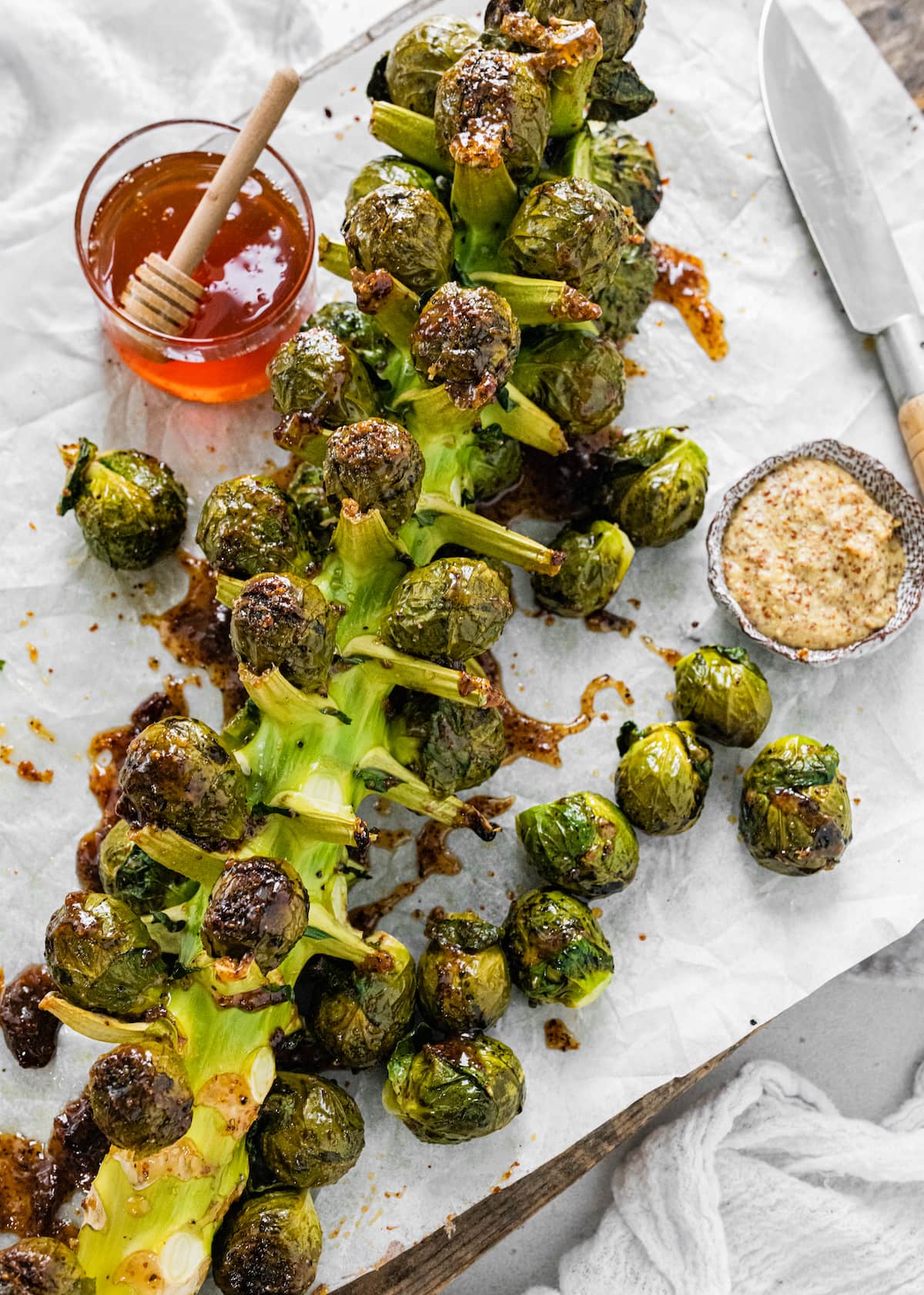 Roasted Brussels Sprouts on the Stalk on parchment paper on a cutting board near a small bowl of mustard, a small container of honey, and a knife.