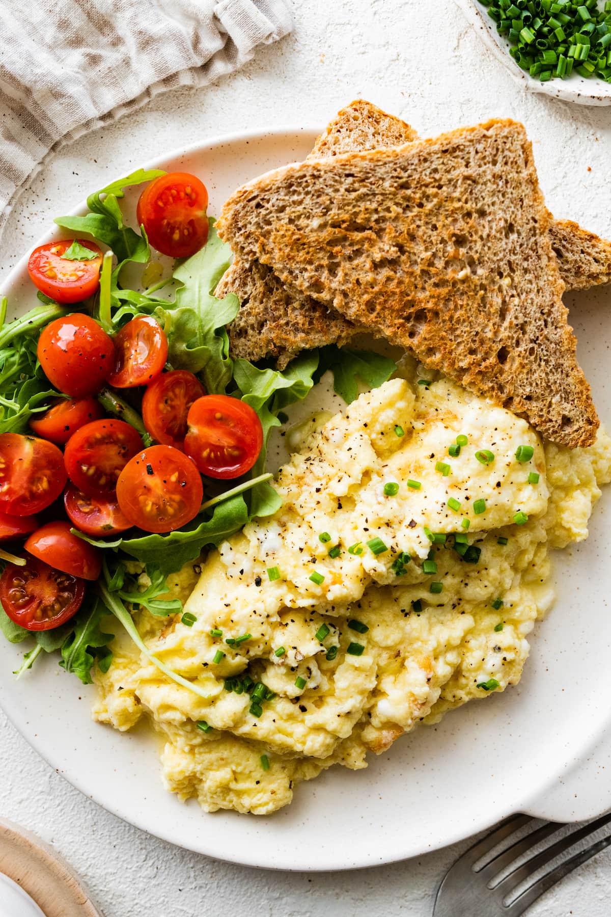 Cottage cheese eggs garnished with chives on a plate with toast and a salad with greens and cherry tomatoes.