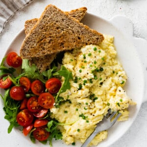 Cottage cheese eggs garnished with chives on a plate with toast and a salad with greens and cherry tomatoes.