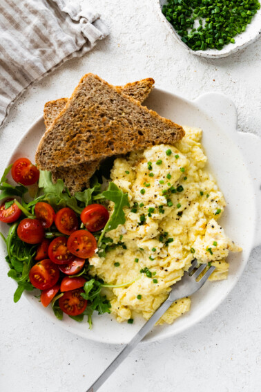 Cottage cheese eggs garnished with chives on a plate with toast and a salad with greens and cherry tomatoes.