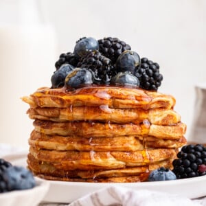 A large stack of cottage cheese pancakes on a plate with maple syrup topped with fresh blueberries and blackberries.