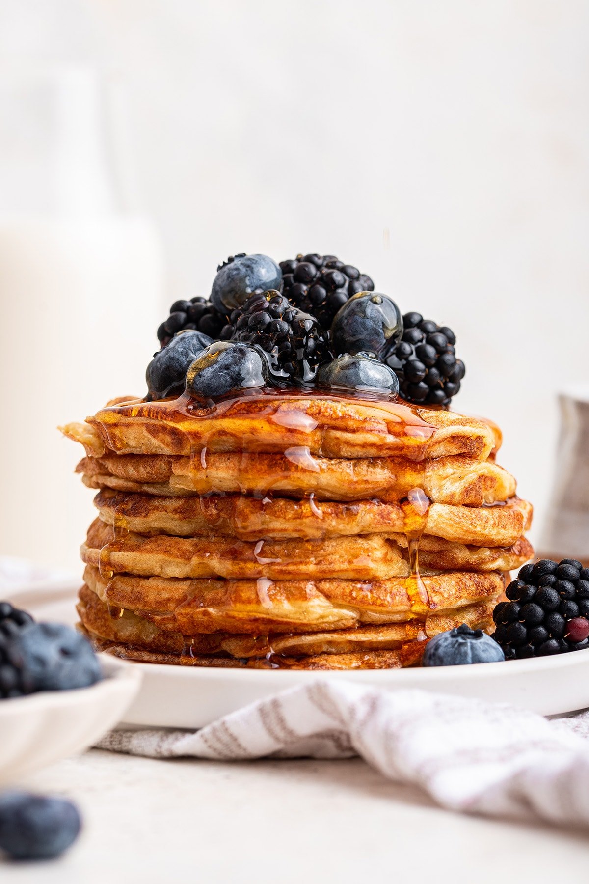 A large stack of cottage cheese pancakes on a plate with maple syrup topped with fresh blueberries and blackberries.
