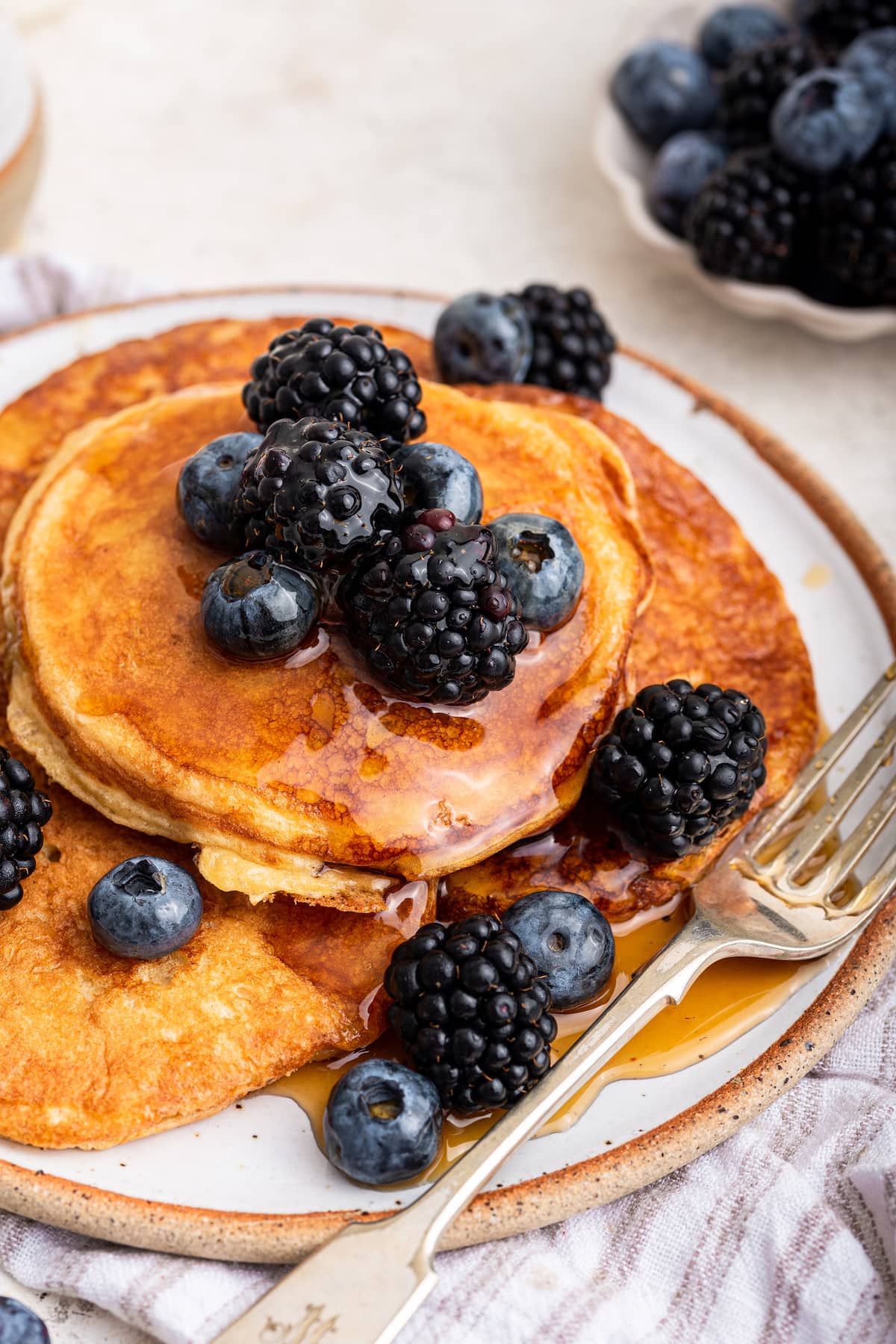 Multiple cottage cheese pancakes on a plate with a fork. The pancakes are topped off with maple syrup and fresh berries.