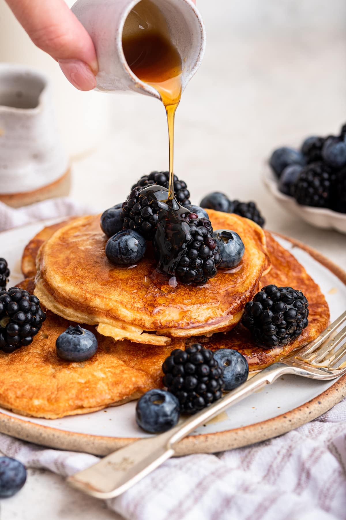 Maple syrup being poured over a stack of cottage cheese pancakes that are topped off with fresh blueberries and blackberries.
