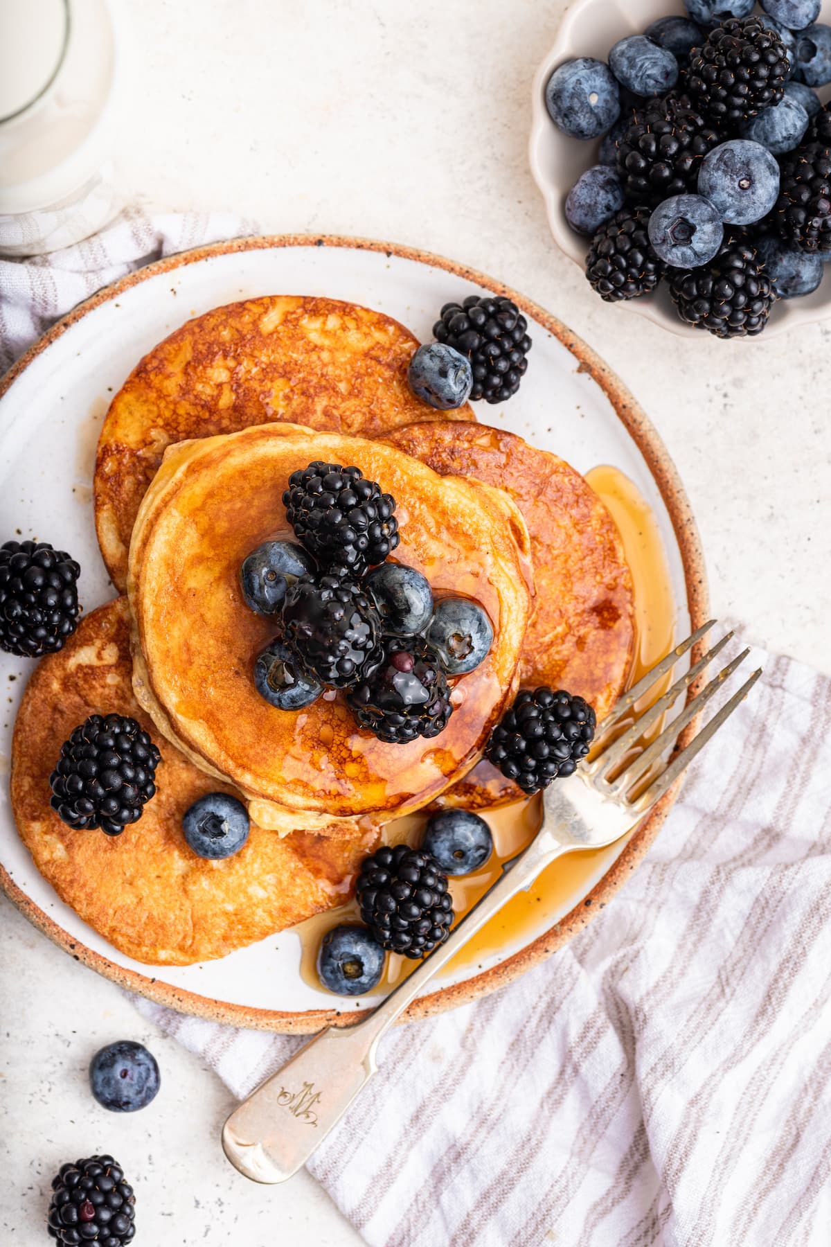Multiple cottage cheese pancakes on a plate with a fork. The pancakes are topped off with maple syrup and fresh berries.