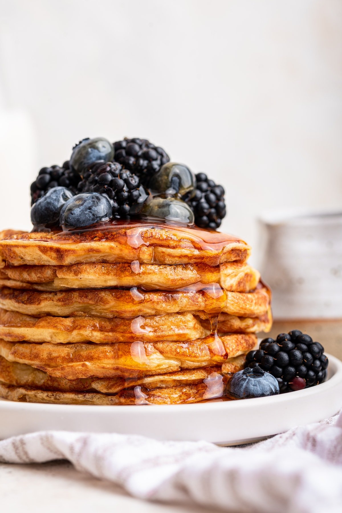 A large stack of cottage cheese pancakes on a plate with maple syrup topped with fresh blueberries and blackberries.