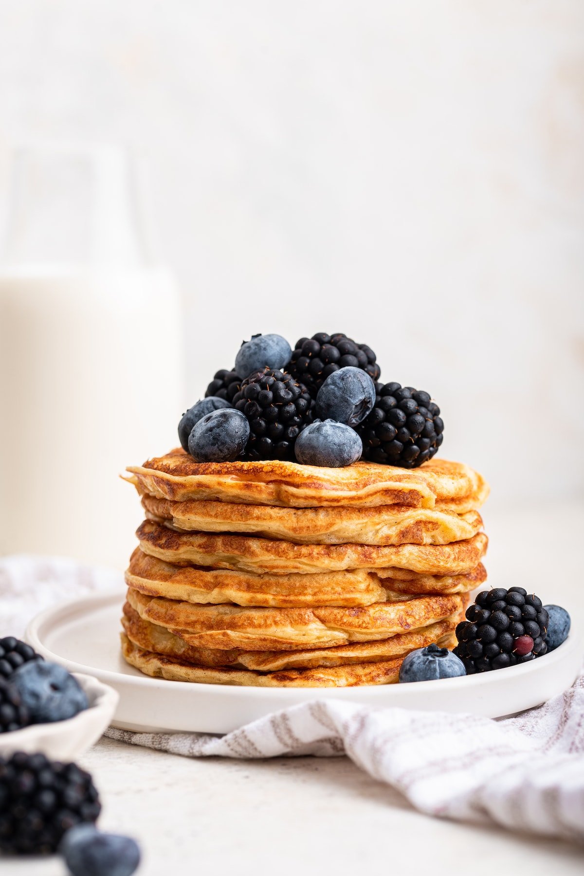 A large stack of cottage cheese pancakes on a plate topped with fresh blueberries and blackberries.