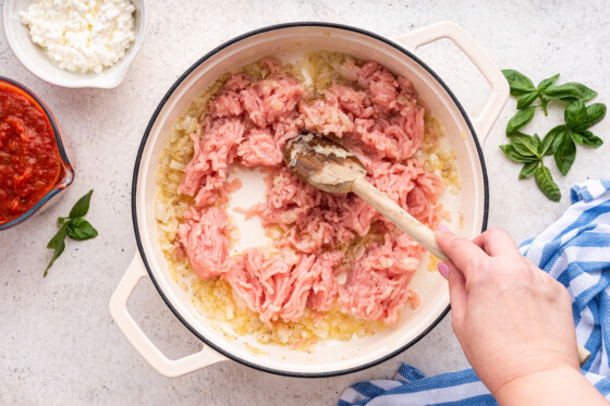 A woman's hand using a wooden spoon to break up ground turkey in a large dutch oven with garlic, onion, and seasonings.