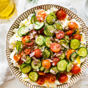 A cottage cheese salad with cucumber, tomatoes, seasoning, and fresh herbs in a large bowl.