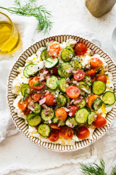 A cottage cheese salad with cucumber, tomatoes, seasoning, and fresh herbs in a large bowl.