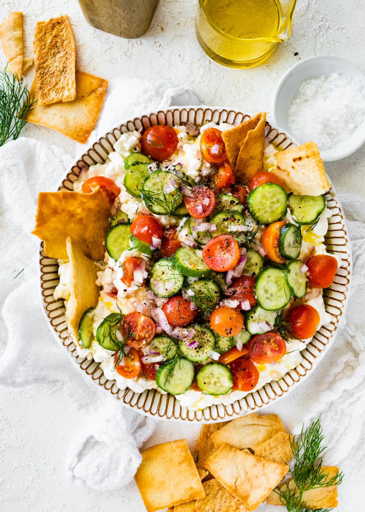 A cottage cheese salad with cucumber, tomatoes, seasoning, and fresh herbs in a large bowl with pita chips on the side.