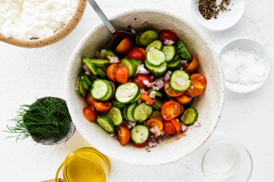 Sliced cucumber, tomatoes, and diced red onion in a large bowl with a metal spoon.