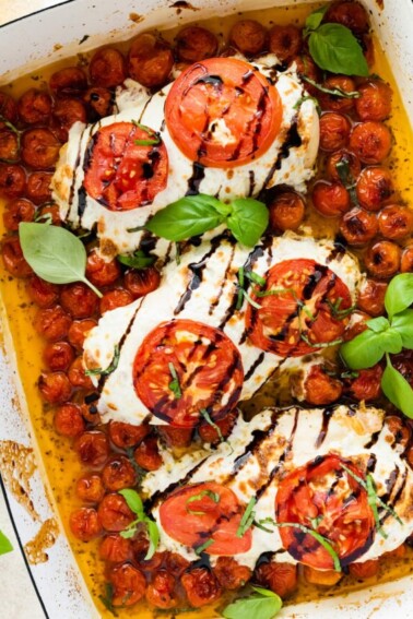 Three chicken breasts covered in mozzarella cheese, tomato slices, and balsamic glaze in a rectangular baking dish with cherry tomatoes and fresh basil.