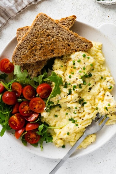 Cottage cheese eggs garnished with chives on a plate with toast and a salad with greens and cherry tomatoes.