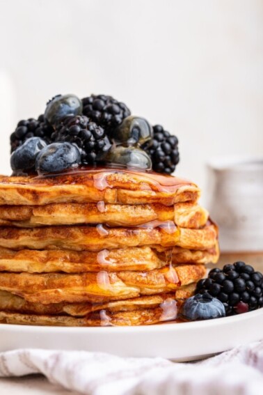 A large stack of cottage cheese pancakes on a plate with maple syrup topped with fresh blueberries and blackberries.