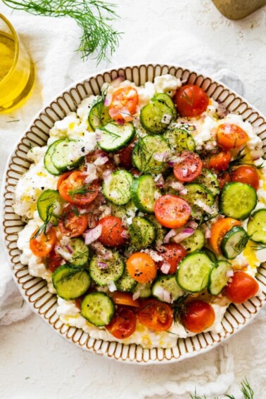 A cottage cheese salad with cucumber, tomatoes, seasoning, and fresh herbs in a large bowl.