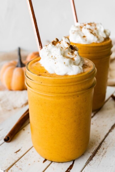 Two pumpkin protein shakes in glass mason jars with a metal straw, topped with whipped cream and ground cinnamon.