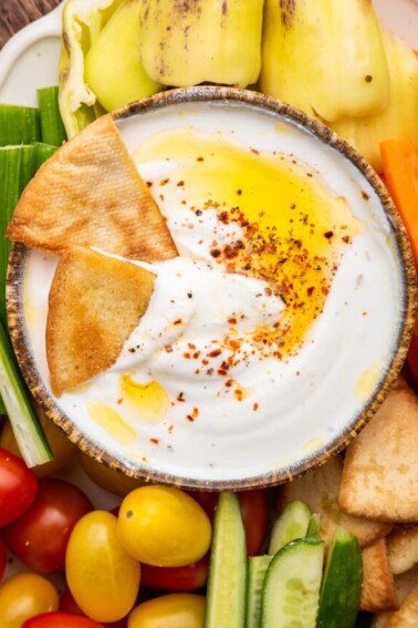 Whipped cottage cheese in a small dipping bowl topped with olive oil and crushed red pepper. Around the bowl are tomatoes, cucumbers, carrots, peppers, and pita chips.