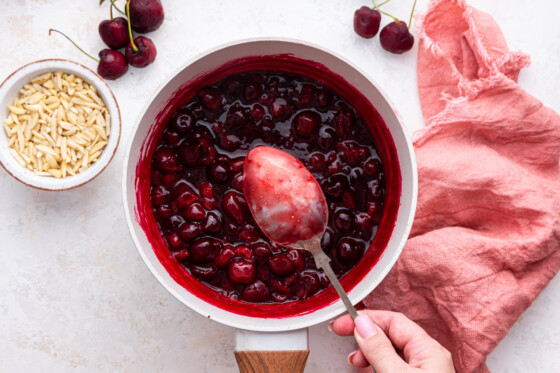 A womans hand hold a spoon over a saucepan of melted cherries.