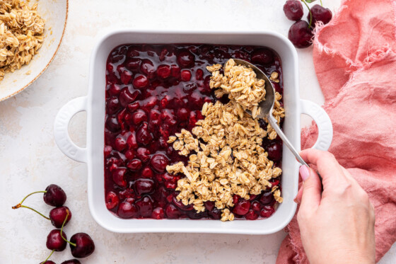A woman's hand uses a spoon to spread the oat crisp over the cherries in a large, square, baking dish.