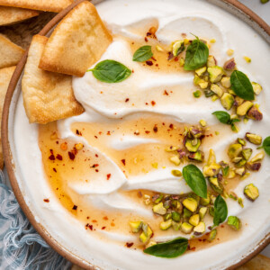 Hot honey whipped cottage cheese in a shallow bowl topped with with a drizzle of hot honey, olive oil, crack of black pepper, fresh basil and chopped pistachios. There are two pita chips in the bowl.