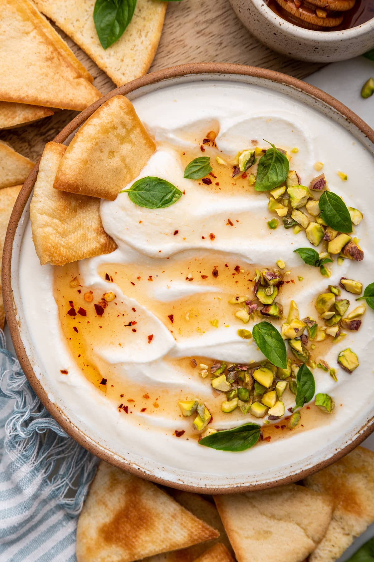 Hot honey whipped cottage cheese in a shallow bowl topped with with a drizzle of hot honey, olive oil, crack of black pepper, fresh basil and chopped pistachios. There are two pita chips in the bowl.