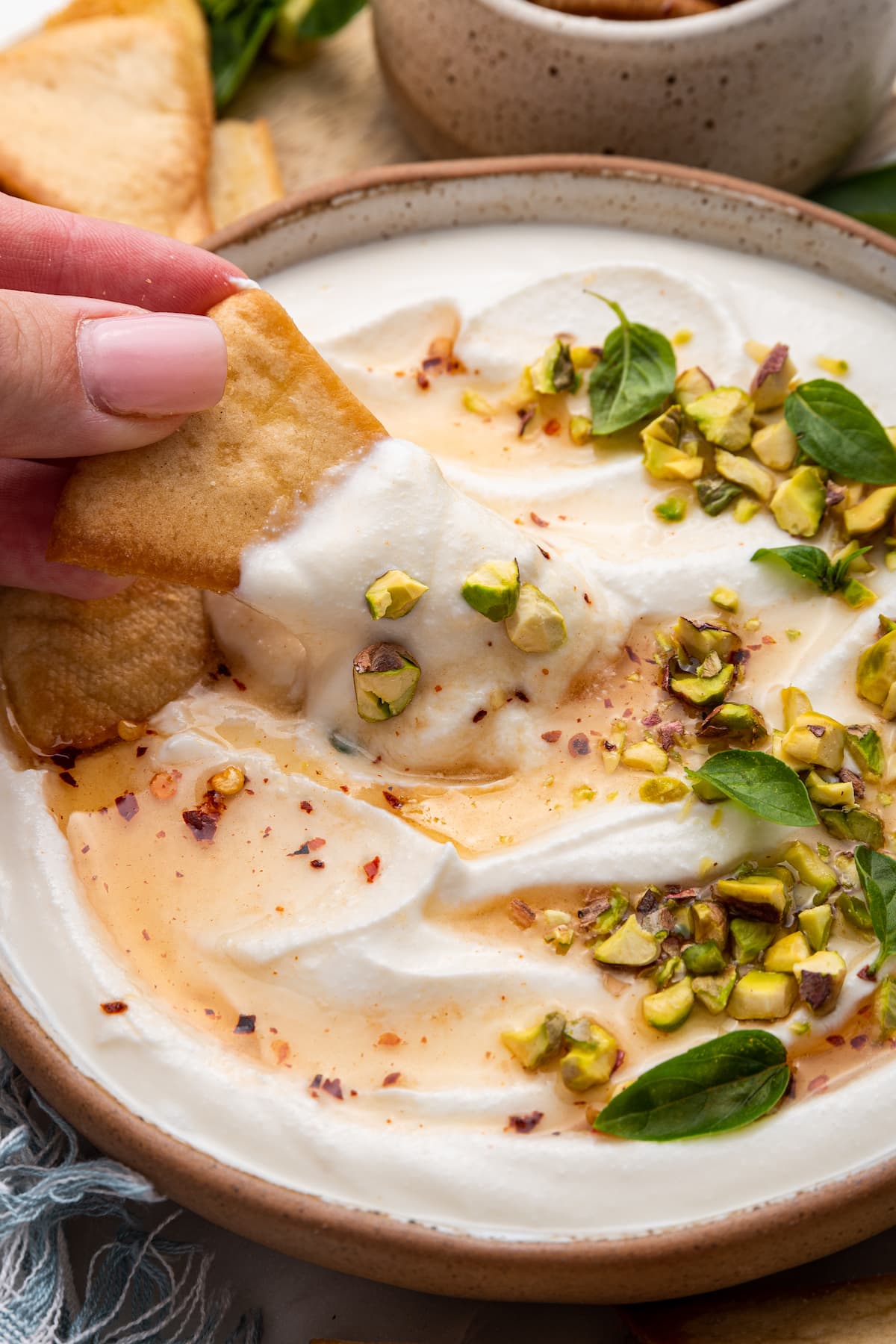 A woman's hand dipping a pita chip in hot honey whipped cottage cheese in a shallow bowl topped with with a drizzle of hot honey, olive oil, crack of black pepper, fresh basil and chopped pistachios.