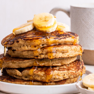 Five overnight oatmeal pancakes stacked on one another on a small plate and topped with banana slices with maple syrup dripping down from each pancake.