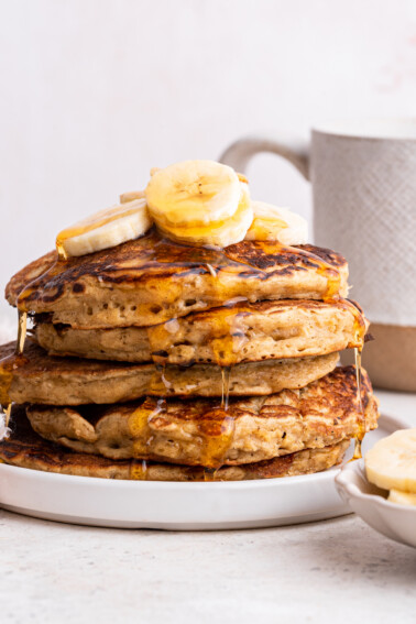 Five overnight oatmeal pancakes stacked on one another on a small plate and topped with banana slices with maple syrup dripping down from each pancake.