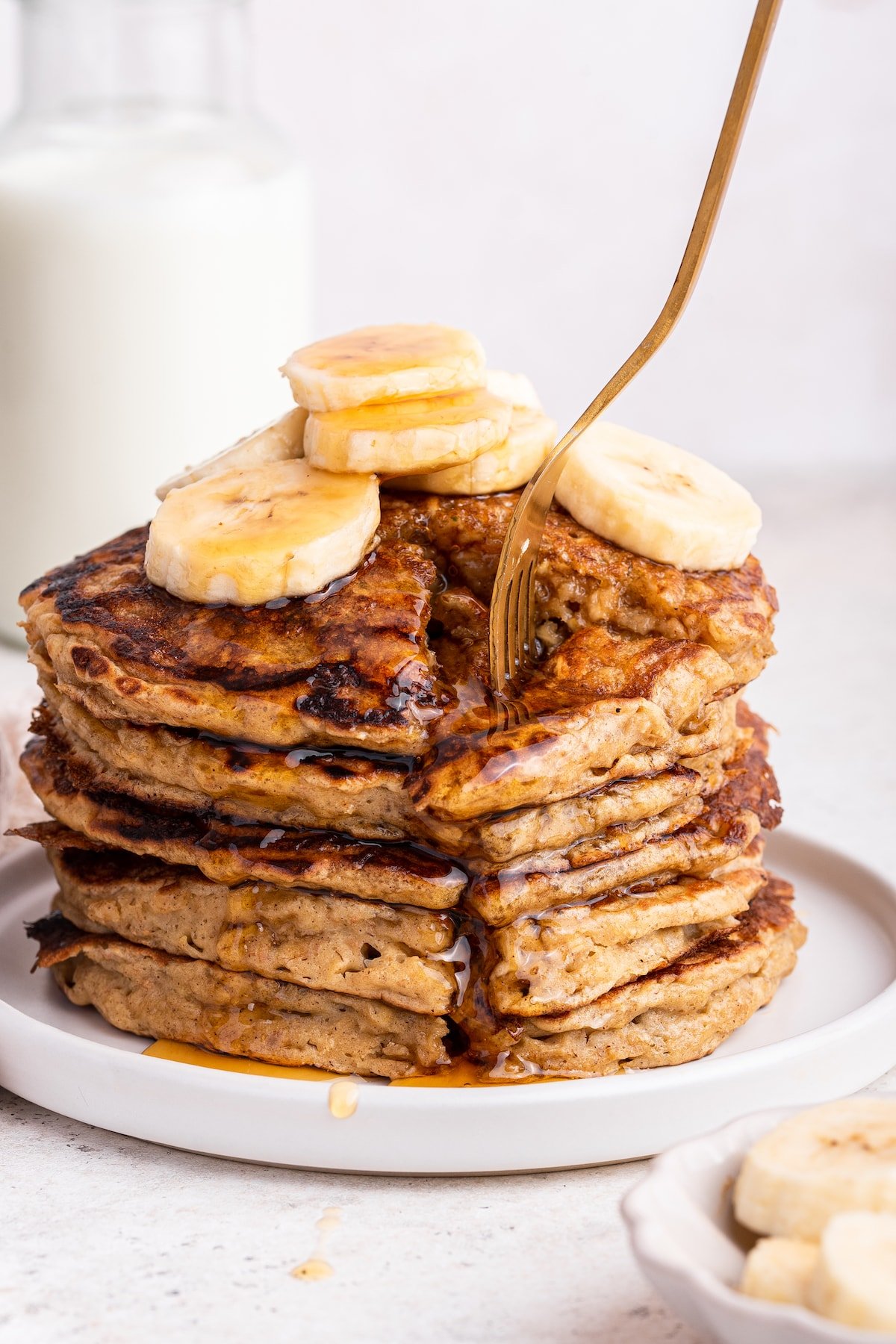 A fork digging into a large portion of a stack of multiple overnight oatmeal pancakes that are topped with banana slices and maple syrup.