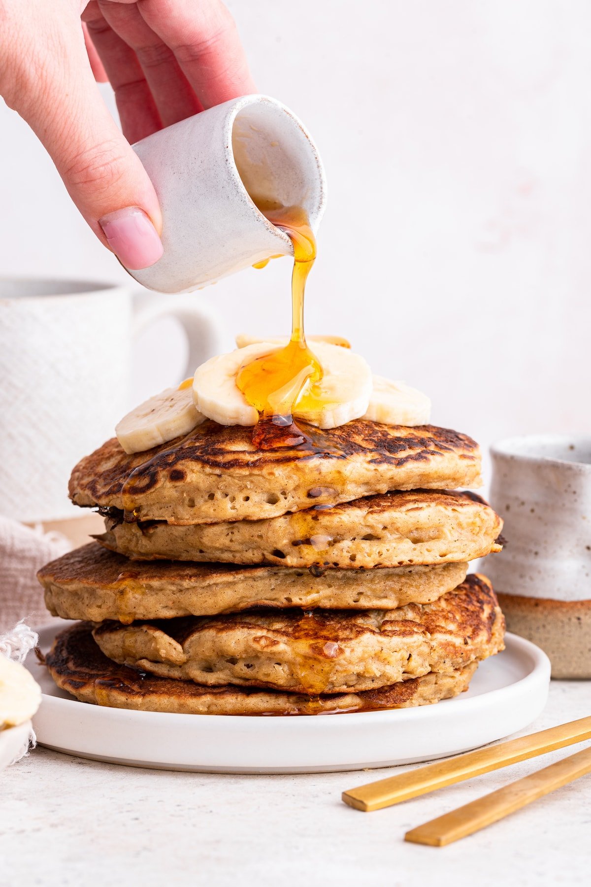 Maple syrup being poured over a stack of multiple overnight oatmeal pancakes that are topped with banana slices.