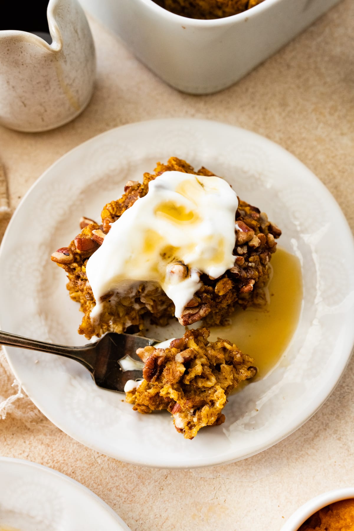 A serving of pumpkin baked oatmeal on a small white plate with fork taking a small piece from it. The slice has a whipped cream and maple syrup topping.