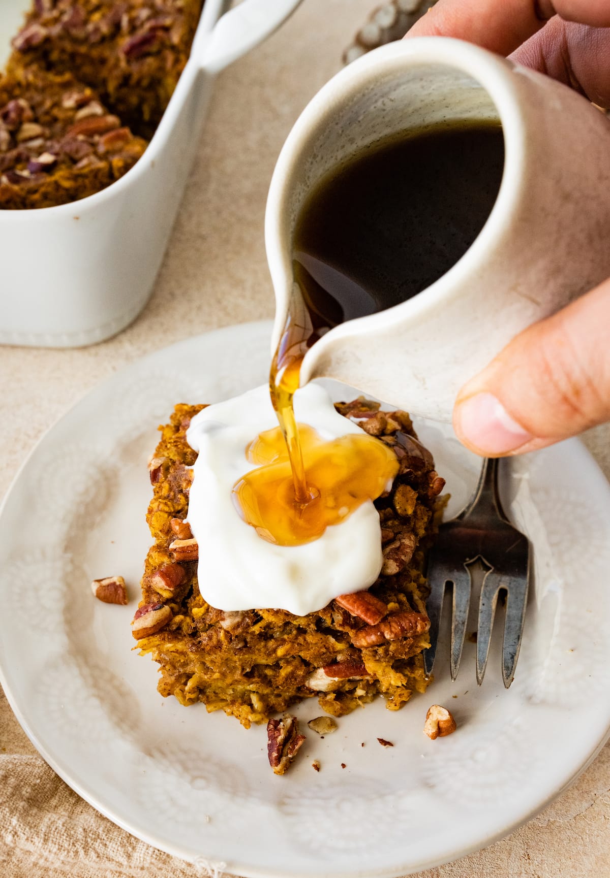 Maple syrup being poured over a slice of pumpkin baked oatmeal that is topped with whipped cream.