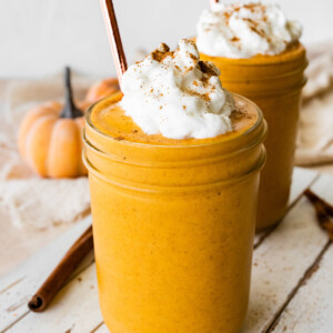 Two pumpkin protein shakes in glass mason jars with a metal straw, topped with whipped cream and ground cinnamon.