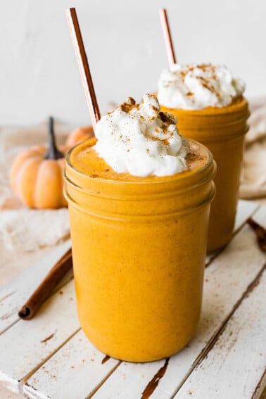 Two pumpkin protein shakes in glass mason jars with a metal straw, topped with whipped cream and ground cinnamon.