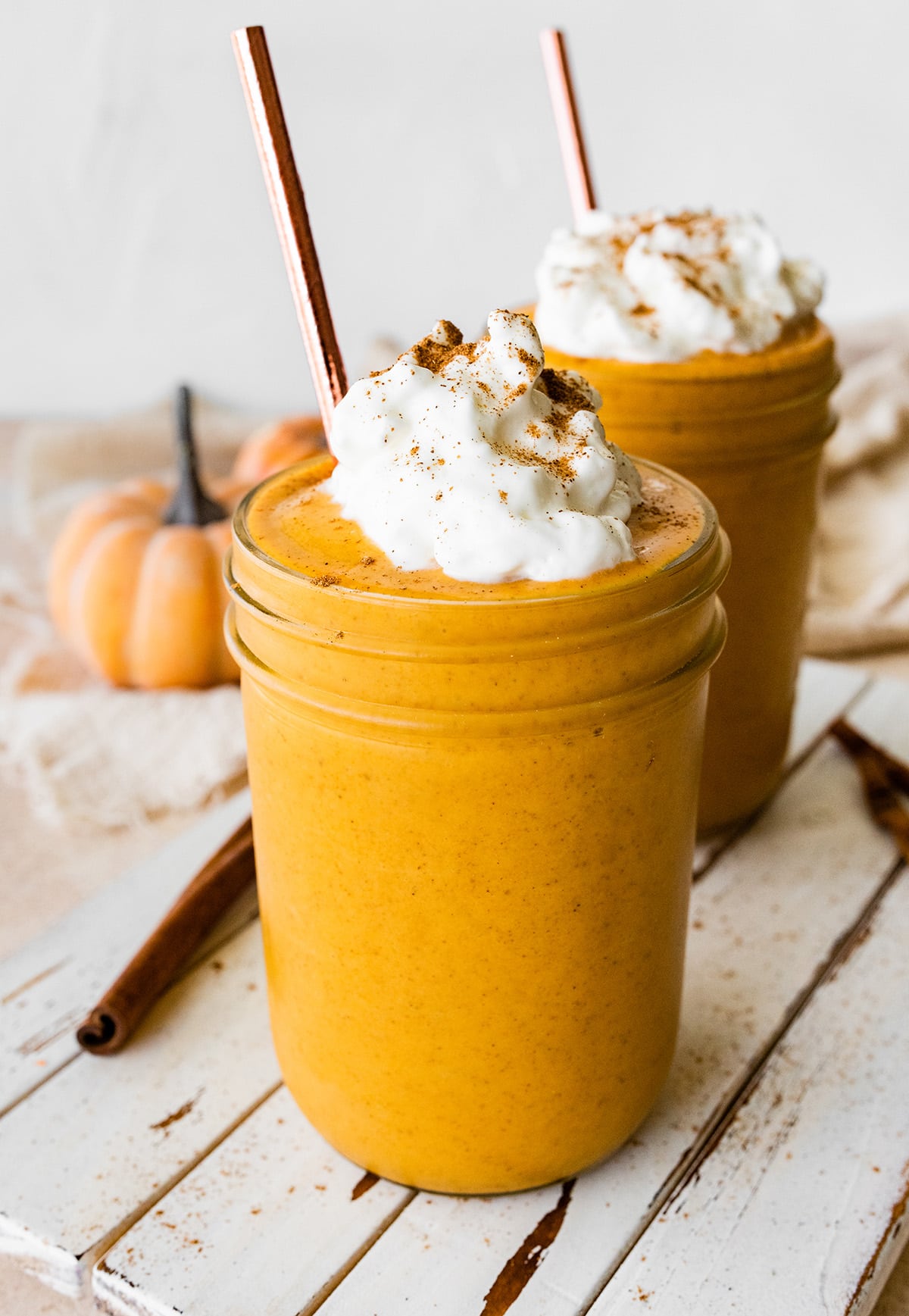 Two pumpkin protein shakes in glass mason jars with a metal straw, topped with whipped cream and ground cinnamon.