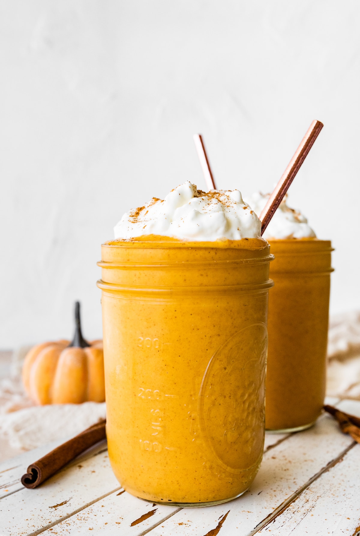 Two pumpkin protein shakes in glass mason jars with a metal straw, topped with whipped cream and ground cinnamon.