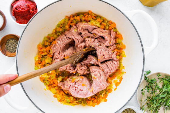 A woman's hand uses a wooden spoon to break up the ground turkey into the vegetables in a large dutch oven.