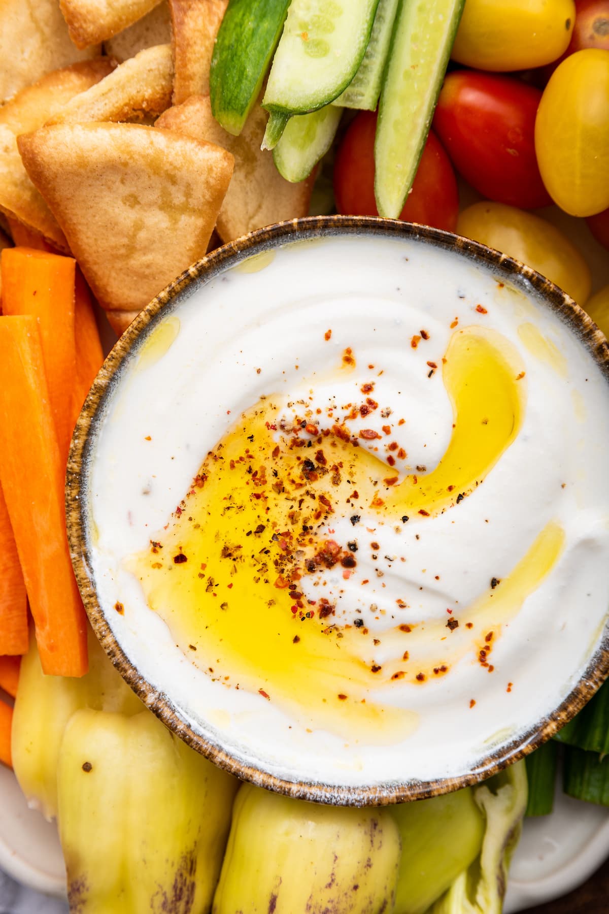 Whipped cottage cheese in a small dipping bowl topped with olive oil and crushed red pepper. Around the bowl are tomatoes, cucumbers, carrots, peppers, and pita chips.