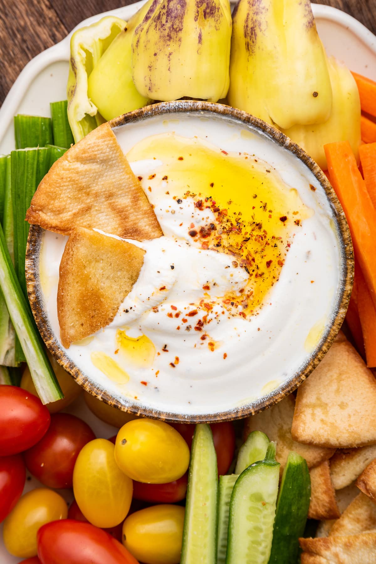 Whipped cottage cheese in a small dipping bowl topped with olive oil and crushed red pepper. Around the bowl are tomatoes, cucumbers, carrots, peppers, and pita chips.