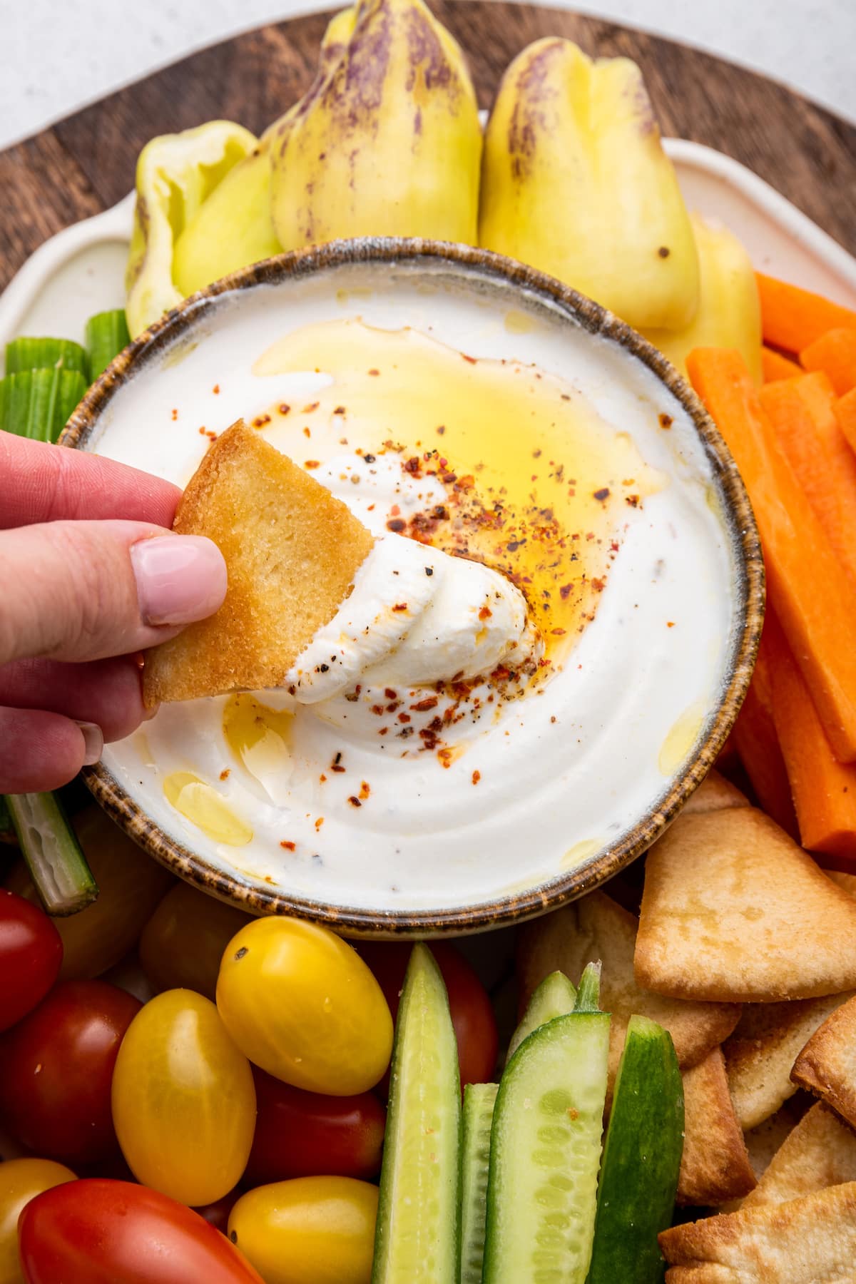 A woman's hand dipping a pita chip in a small bowl of whipped cottage cheese topped with olive oil and crushed red pepper. Around the bowl are tomatoes, cucumbers, carrots, peppers, and pita chips.