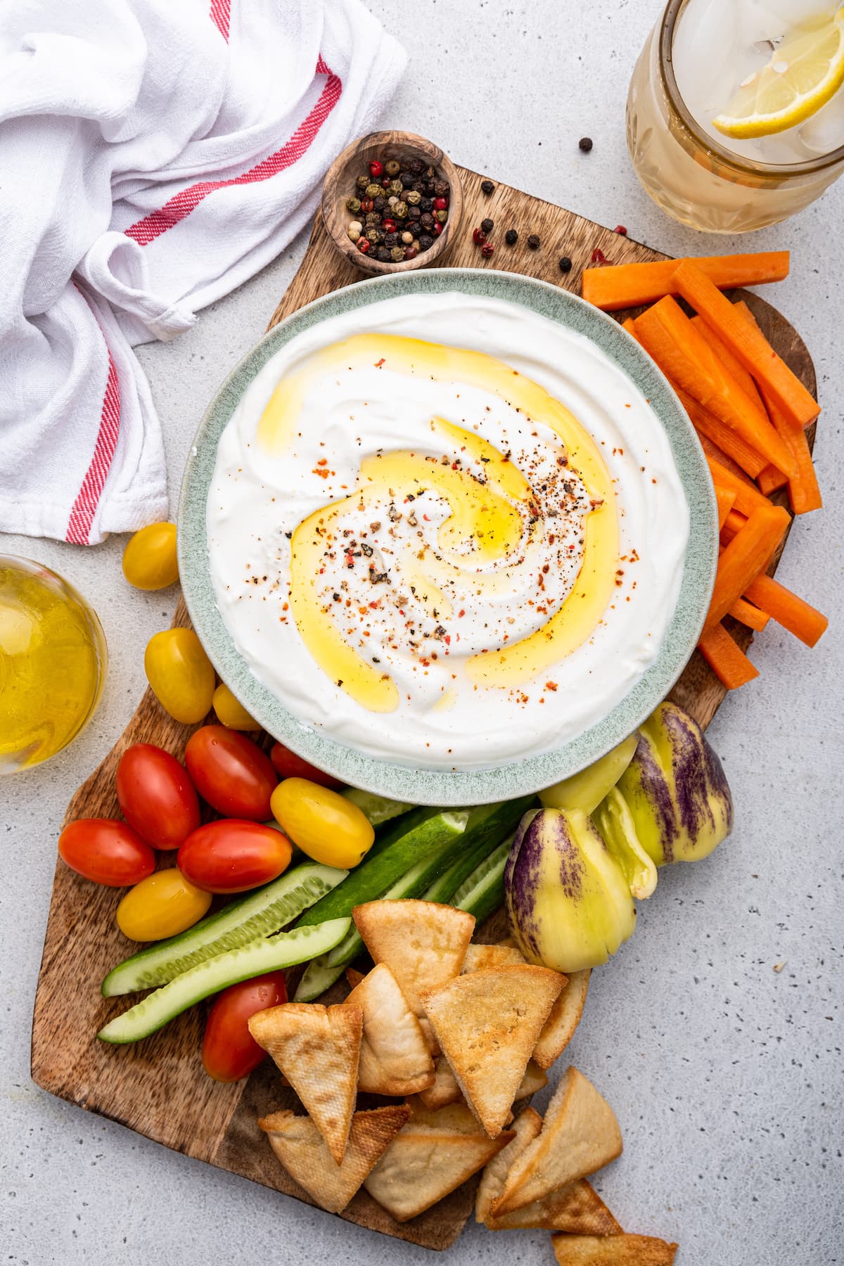 Whipped cottage cheese on a plate topped with olive oil and crushed red pepper. The plate is on a wooden cutting board along with tomatoes, cucumbers, pita chips, peppers, and carrots.