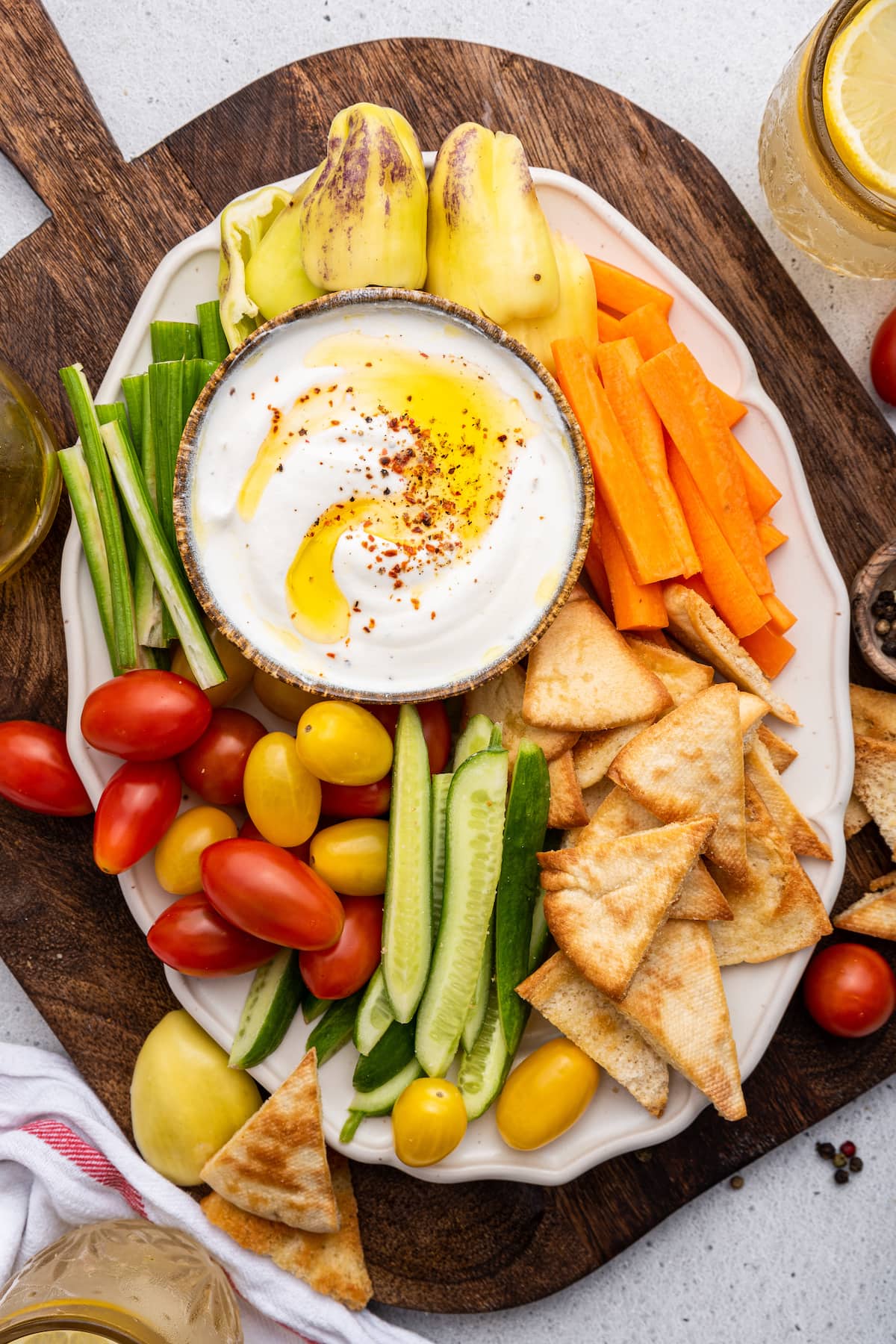 Whipped cottage cheese in a small dipping bowl that is on a serving plate that is on a wooden cutting board. Around the small bowl of whipped cottage cheese are vegetables and chips that include tomatoes, carrots, cucumbers, peppers, and pita chips.