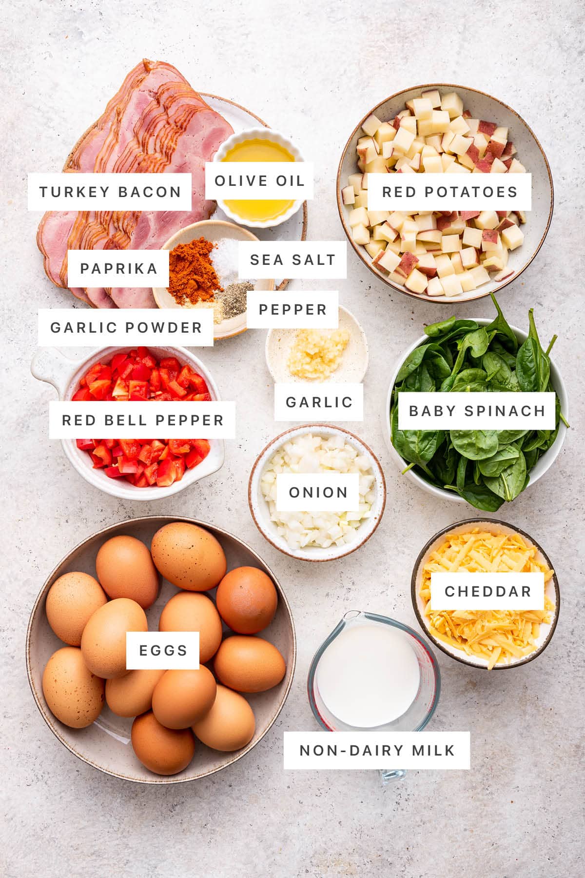 Ingredients measured out to make Make-Ahead Breakfast Casserole: turkey bacon, olive oil, paprika, sea salt, pepper, garlic powder, garlic, red potatoes, red bell pepper, onion, baby spinach, eggs, non-dairy milk and cheddar.